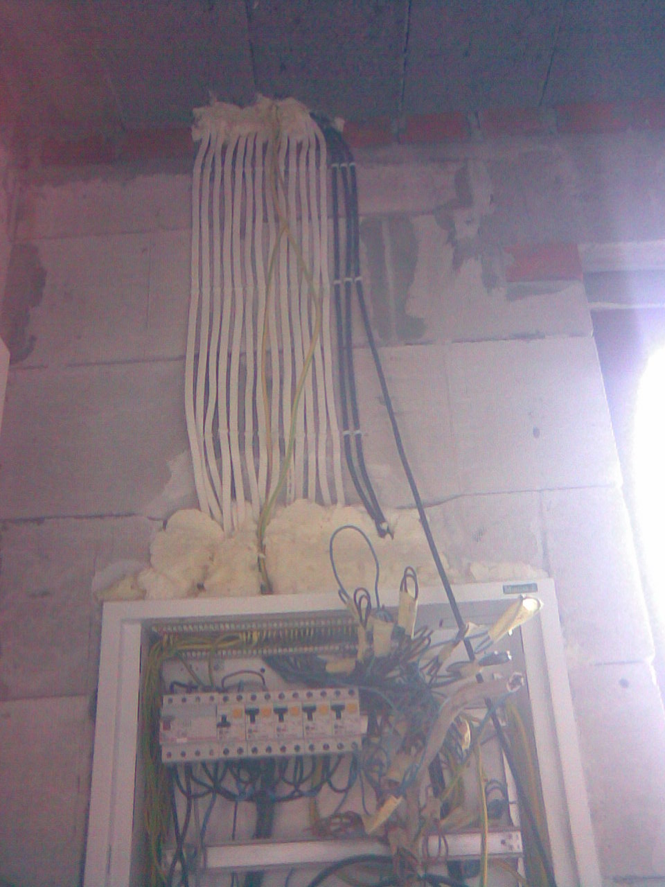 Rozdzielnica elektryczna w trakcie montażu w surowym wnętrzu, kable zabezpieczone pianką montażową, widoczne przewody i wyłączniki.